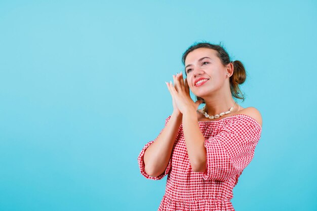 La chica sonriente está mirando hacia arriba tomándose de la mano cerca de la cara con fondo azul.