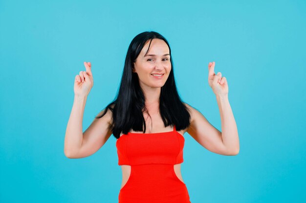 Chica sonriente está mirando a la cámara levantando los dedos cruzados sobre fondo azul.