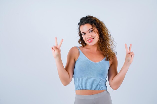 Chica sonriente está mirando a la cámara mostrando dos gestos sobre fondo blanco.