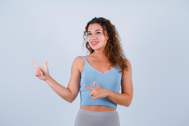 La chica sonriente está mirando hacia otro lado señalando a la izquierda con los dedos índices sobre fondo blanco.