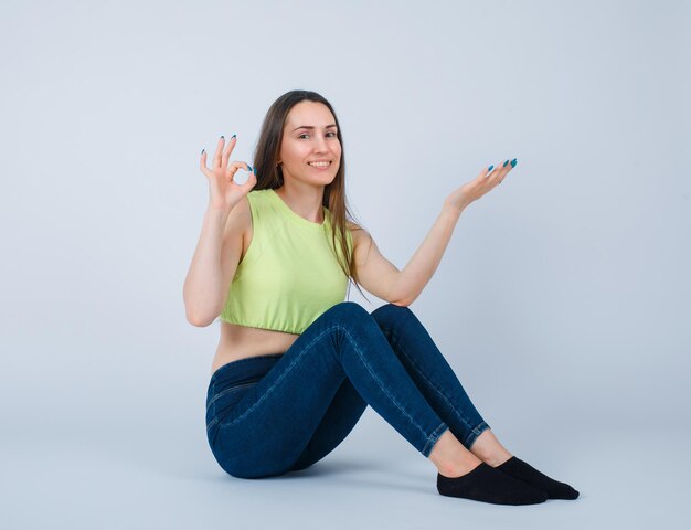 La chica sonriente está mostrando un gesto correcto al sentarse en el suelo sobre fondo blanco.