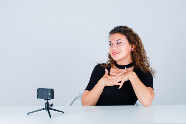 Chica sonriente está posando para su mini cámara tomándose de la mano en el pecho sobre fondo blanco.