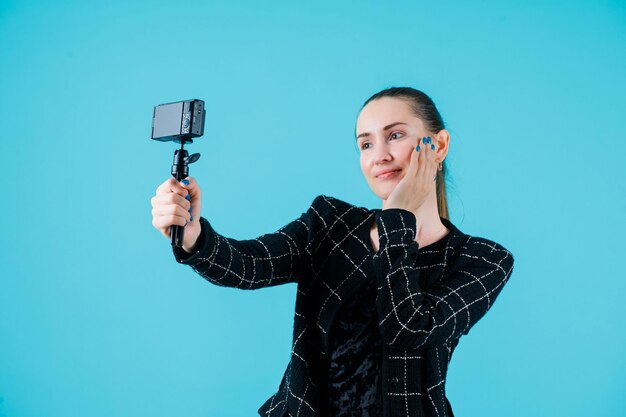 Chica sonriente se está tomando selfie poniendo la mano en la mejilla sobre fondo azul.