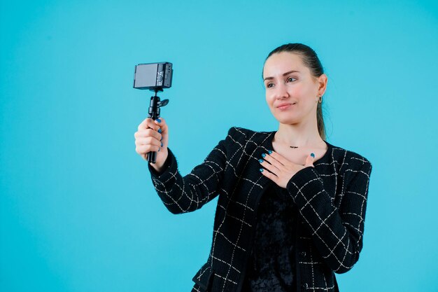 Chica sonriente se está tomando selfie con su cámara sosteniendo la mano en el pecho sobre fondo azul.
