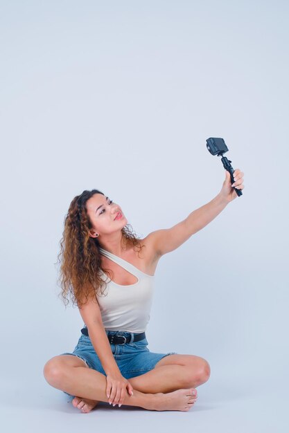 Una chica sonriente se está tomando una selfie con su mini cámara sentada en el suelo con fondo blanco