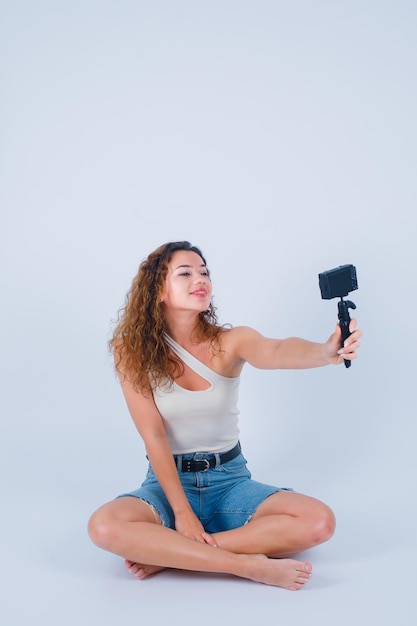 Una chica sonriente se está tomando una selfie con su mini cámara sentada en el suelo con fondo blanco