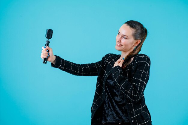 La chica sonriente se está tomando una selfie con su mini cámara sosteniéndola en el pecho con fondo azul.