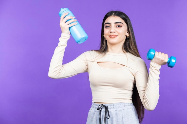 Chica sonriente haciendo ejercicio y sosteniendo una botella de agua Chica joven en forma de pie sobre fondo púrpura Foto de alta calidad