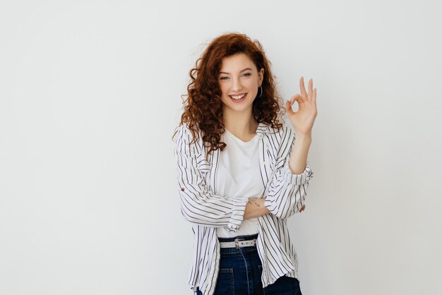 Una chica sonriente impresionada con el pelo rojo muestra el signo OK OK y se ve asombrada alabanza oferta promocional genial hacer cumplido mostrar cero gesto sobre fondo blanco