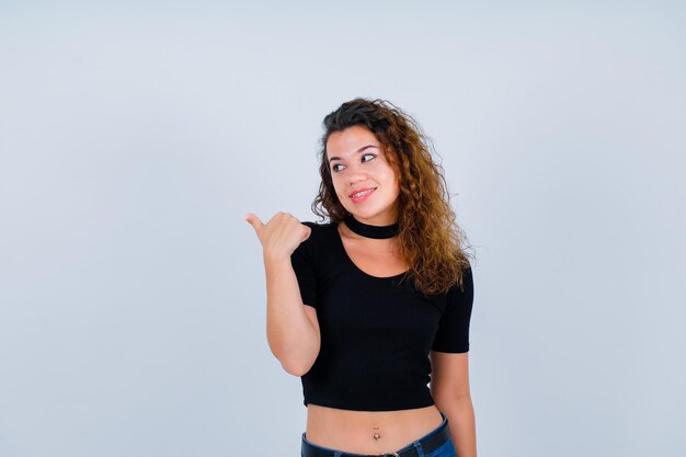 La chica sonriente mira a la izquierda apuntando hacia atrás con el pulgar en el fondo blanco