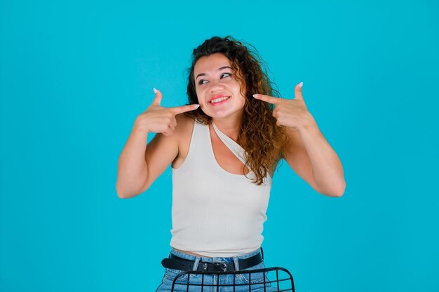 La chica sonriente mira a la izquierda señalando su sonrisa con los dedos índices sobre fondo azul.