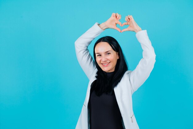 La chica sonriente muestra un gesto de corazón levantando las manos por encima de la cabeza con fondo azul.