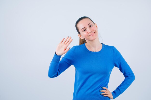 La chica sonriente muestra un gesto de hola y pone la otra mano en la cintura con fondo blanco