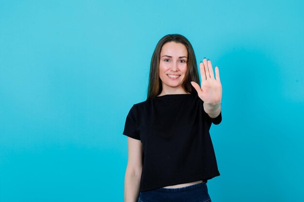La chica sonriente muestra un gesto de parada extendiendo la mano a la cámara con fondo azul.