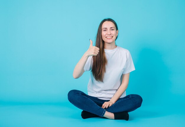 La chica sonriente muestra un gesto perfecto al sentarse en el suelo con fondo azul.