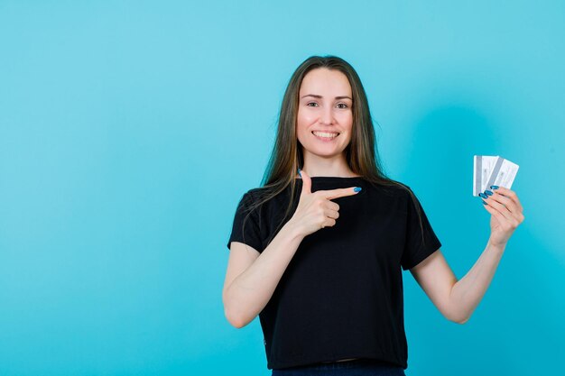 Chica sonriente muestra tarjetas de crédito en la mano con el dedo índice sobre fondo azul.