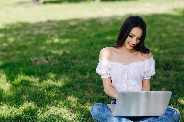 Chica sonriente sentada en un parque escribiendo en su portátil