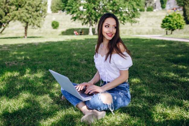 Chica sonriente sentada en un parque escribiendo en su portátil