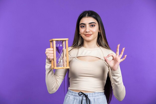 Chica sonriente sosteniendo reloj de arena y gesto OK con la mano