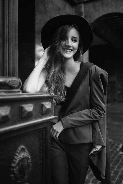 Chica sonriente sosteniendo un sombrero de pie en la calle. Modelo de moda en la ciudad. Fotografía de moda en blanco y negro. Mujer elegante con siut y sombrero.