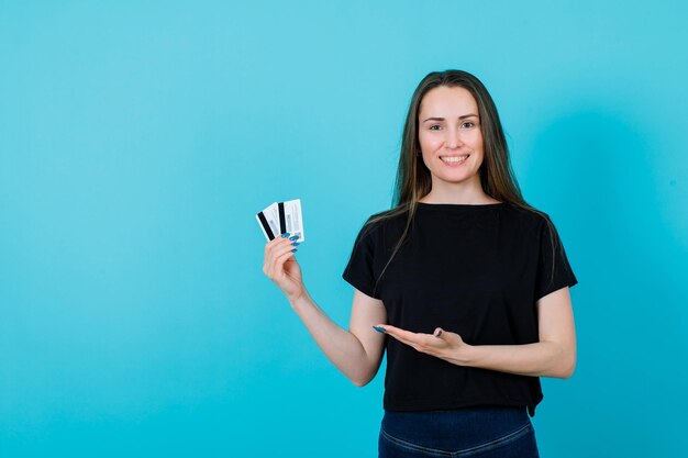 La chica sonriente tiene tarjetas de crédito y las muestra con la otra mano en el fondo azul