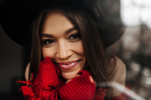 Chica con una sonrisa encantadora mira a la cámara posando con un sombrero de ala ancha y una chaqueta brillante tejida