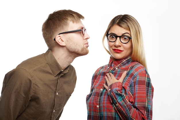 Chica sorprendida con camisa a cuadros de estilo y gafas ovaladas sacando los ojos, sosteniendo la mano en su pecho, sintiéndose aterrorizada mientras un chico nerd la va a besar, haciendo pucheros y cerrando los ojos.