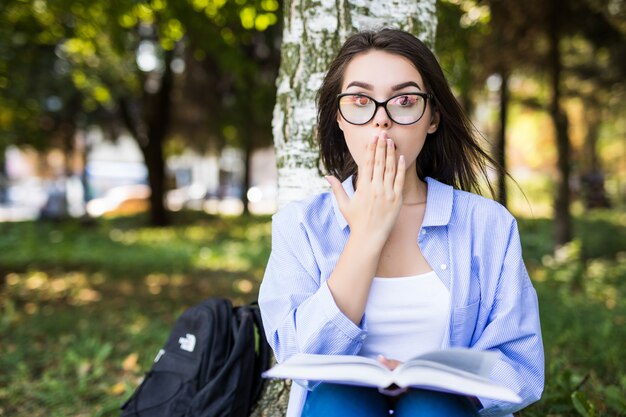 Chica sorprendida en chaqueta de jeans y gafas lee el libro contra el parque verde de verano.