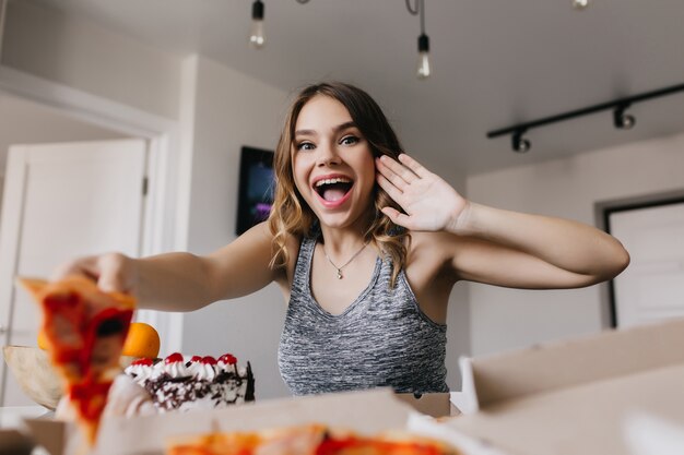 Chica sorprendida comiendo pizza con tomates. Foto interior de mujer blanca bonita disfrutando de comida rápida en la mañana del fin de semana.