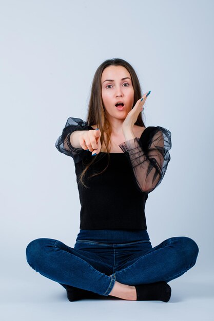 Chica sorprendida está extendiendo el dedo índice a la cámara sentándose en el suelo sobre fondo blanco.