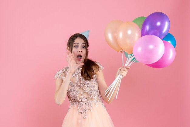 Chica sorprendida con gorra de fiesta sosteniendo globos saludando a alguien en rosa