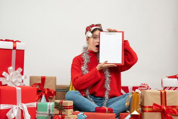 Chica sorprendida con gorro de Papá Noel que cubre su ojo con documento sentado alrededor presenta en blanco