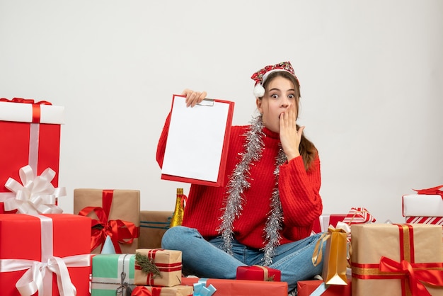 Chica sorprendida con gorro de Papá Noel sosteniendo documentos sentados alrededor presenta en blanco