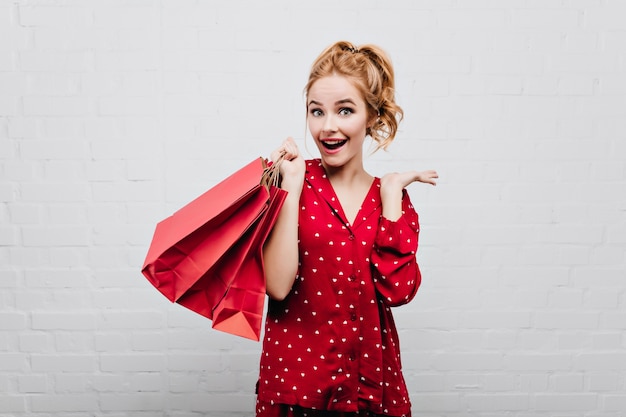 Chica sorprendida de ojos azules en ropa de dormir de algodón rojo posando con una sonrisa en la pared blanca Maravillosa dama en pijama divirtiéndose en la mañana de cumpleaños, con bolsa de regalo.