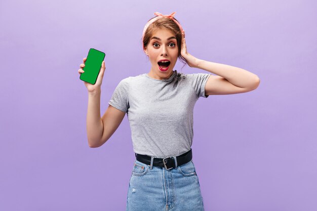 Chica sorprendida posa con teléfono sobre fondo morado. Conmocionada mujer moderna en camiseta ligera y falda con cinturón sostiene smartphone.