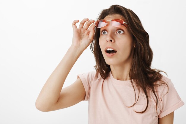 Chica sorprendida y sorprendida con gafas de sol posando contra la pared blanca