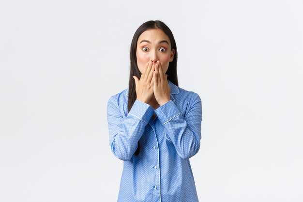 Chica sorprendida sorprendida presencia algo horrible y sorprendente. Mujer asiática en pijama azul reacciona asombrada, cubre la boca con la mano con asombro y mira a la cámara sin palabras sobre fondo blanco.