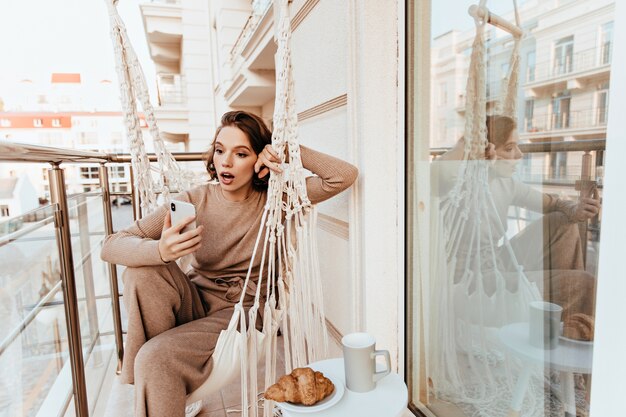 Chica sorprendida en suéter marrón posando con teléfono en el balcón. Señora encantadora asombrada almorzando en la terraza.