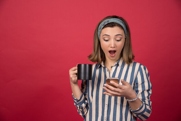 Chica sorprendida con una taza de té comprobando el teléfono móvil