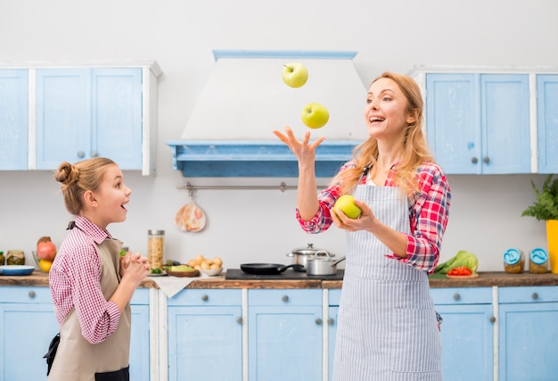 Chica sorpresa mirando a su madre lanzando la manzana verde en el aire en la cocina