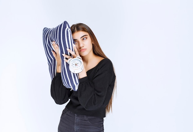 Chica sosteniendo una almohada rayada y mostrando el despertador con una cara triste.