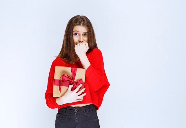 Chica sosteniendo una caja de regalo de cartón envuelto con cinta roja y parece estresada y aterrorizada.