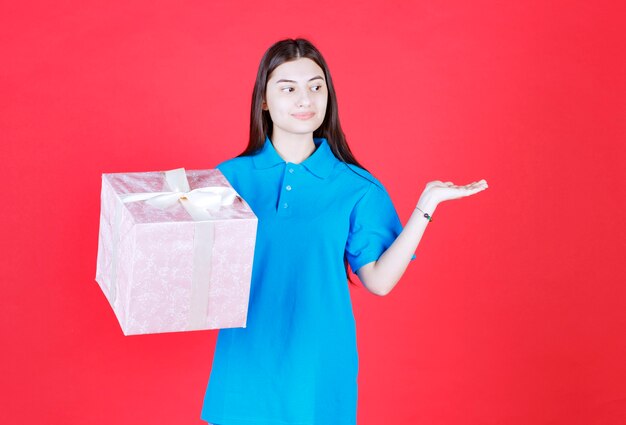 Chica sosteniendo una caja de regalo morada envuelta con cinta blanca e invitando a alguien a presentar el regalo