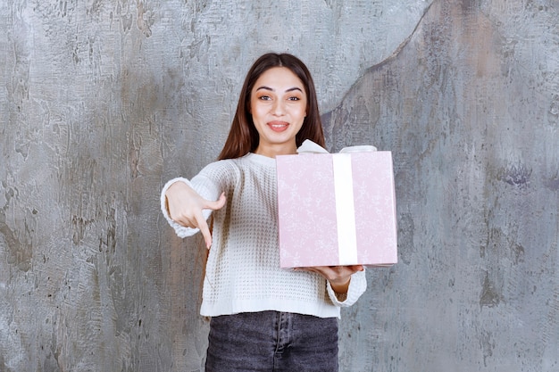 Chica sosteniendo una caja de regalo morada envuelta con cinta blanca e invitando a alguien a presentarla.