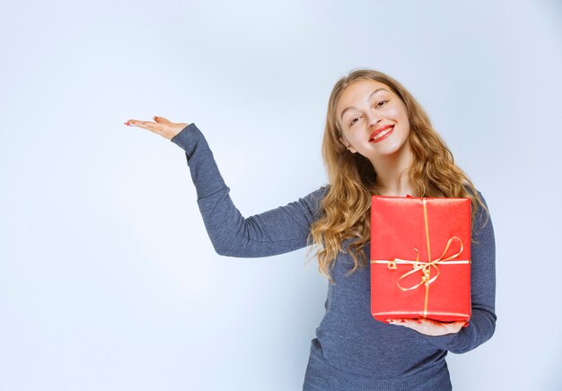Chica sosteniendo una caja de regalo roja y apuntando a alguien de la izquierda.