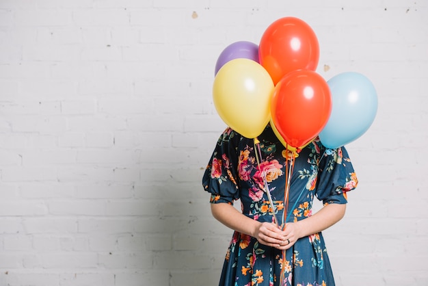 Chica sosteniendo globos de colores frente a su cara de pie contra la pared