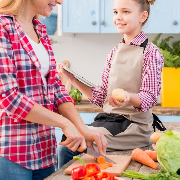 Chica sosteniendo papa y tableta digital en la mano mirando a su madre cortando verduras con un cuchillo