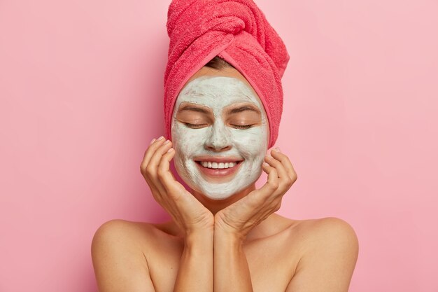 La chica del spa aplica una máscara de arcilla en la cara, mantiene los ojos cerrados, toca las mejillas, disfruta del procedimiento de belleza, refresca la piel, sonríe positivamente aislado en la pared rosa. Relajación, estilo de vida saludable