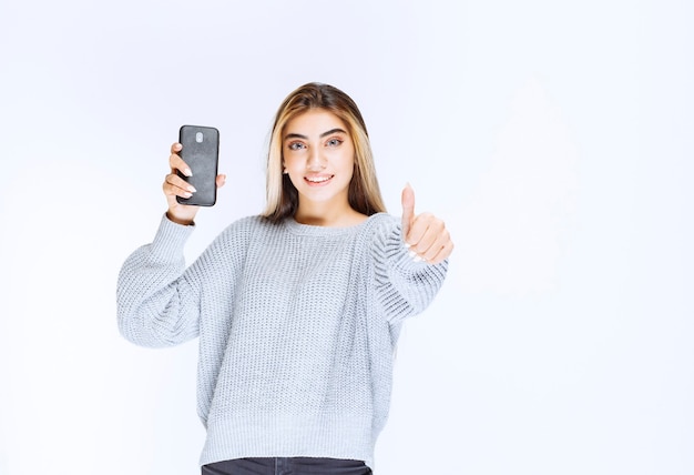 Chica en sudadera gris sosteniendo un teléfono inteligente negro y sintiéndose satisfecha.