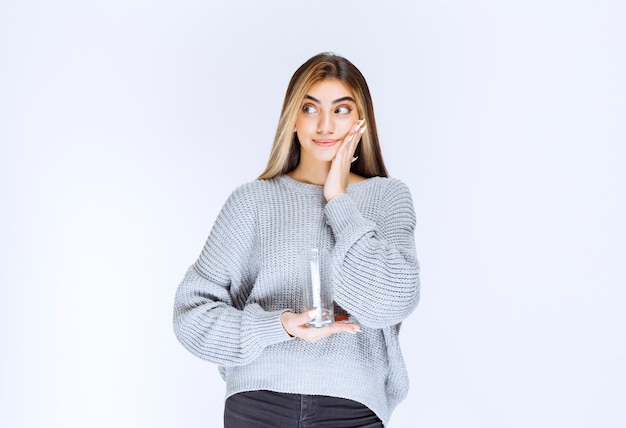 Chica en sudadera gris sosteniendo un vaso de agua pura y haciendo cara de duda.